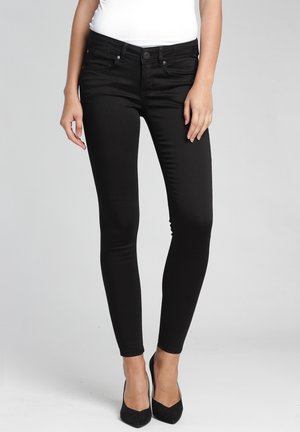 Femme portant un jean skinny noir et des talons noirs, debout avec les mains détendues le long du corps sur un fond gris uni.