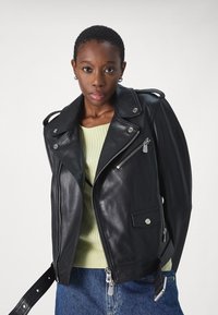 Crna kožna biker jakna sa srebrnim detaljima, džepovima na zatvaranje, asimetričnim prednjim delom i epauletima, složena preko svetlozelenog rebrastog džempera.