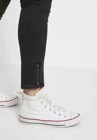 Zapatillas altas blancas con cordones, de material de lona texturizada, combinadas con unos jeans ajustados negros que tienen un detalle de cremallera en el costado del tobillo.