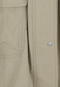 Beige Jacke aus strukturiertem Stoff, mit einer großen Tasche, kontrastierenden Nähten und einem silbernen Druckknopfverschluss.