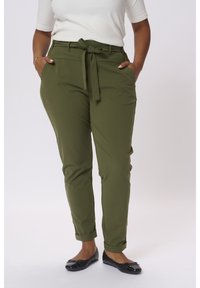 Pantaloni verde oliva a sigaretta con un nodo frontale, tasche laterali e orli arrotolati, abbinati a un top bianco aderente e ballerine nere.