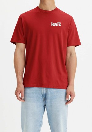 Mand iført en ensfarvet rød Levi's t-shirt og lyseblå jeans, stående med armene afslappet mod en ensfarvet hvid baggrund.