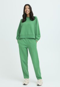 Zelená mikina s uvoľneným strihom a zodpovedajúcimi teplákmi vyrobená z mäkkej tkaniny. Má jednoduchý dizajn s rebrovanými manžetami a bočnými vreckami.