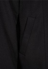 Primer plano de un bolsillo de chaqueta de tela negra con costuras visibles y una abertura en forma de ojal inclinada.