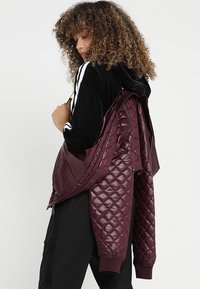 Bordeaux gewatteerde jas van glanzend materiaal met ribgebreide manchetten. Heeft een gelaagd ontwerp met een contrasterende zwarte capuchon en mouwen.
