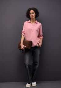 Lichtroze blouse met lange mouwen en een kraag, donkergrijze wijde jeans en een bruine suède clutch, gecombineerd met witte sneakers.