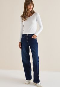 Top blanco de manga larga combinado con jeans de pierna ancha oscuros con cierre de botones, bolsillo lateral y costuras contrastantes.
