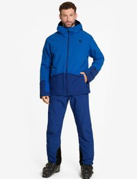 Giacca isolata blu con cappuccio, caratterizzata da un design bicolore. Abbinata a pantaloni blu coordinati. Il materiale appare resistente e impermeabile.