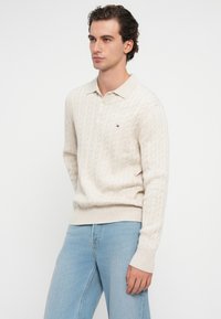 Cremfarbener Poloshirt mit Kabelstrickmuster, ribbelte Bündchen und Saum, mit einem kleinen Logodetail; kombiniert mit hellblauen Jeans.