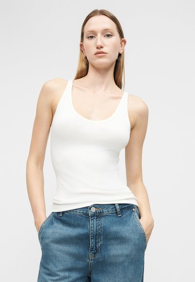 Γυναίκα με μακριά ξανθά μαλλιά που φορά σκουλαρίκια κρίκους, λευκή μπλούζα χωρίς μανίκια και μπλε τζιν, όρθια με τα χέρια στις τσέπες μπροστά σε απλό φόντο.