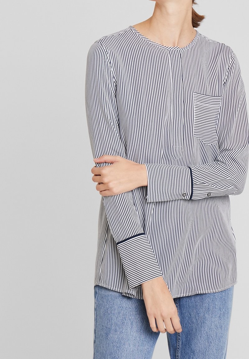 Blusa a maniche lunghe a righe in blu navy e bianco, con scollatura rotonda, taschino sul petto e polsini con bottoni. Tessuto morbido con una texture liscia.