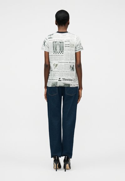 Γυναίκα που φαίνεται από πίσω, φορώντας κοντομάνικο μπλουζάκι με τύπωμα εφημερίδας, σκούρο μπλε τζιν και μαύρα ψηλοτάκουνα παπούτσια σε απλό φόντο.