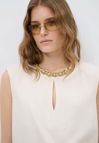 Top color crema con un escote en forma de ojo de cerradura, adornado con un collar de cuentas de oro y cristal. La modelo lleva gafas de sol con tinte amarillo.