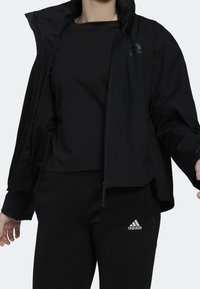 Personne portant un pantalon de survêtement noir Adidas, un t-shirt noir et une veste noire partiellement zippée avec des logos Adidas visibles.