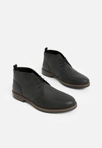 Las botas chukka de cuero negro presentan un acabado suave, diseño de cordones, punta redondeada y una suela intermedia cosida en marrón.
