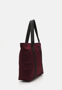 Sac fourre-tout en nylon bordeaux avec des anses noires portant le logo, de forme rectangulaire, texture lisse et design simple. Pas de matériel ou de motifs visibles.