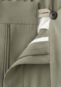 Gros plan sur un pantalon vert olive avec braguette dézippée, ceinture intérieure blanche et fermeture par bouton noir.