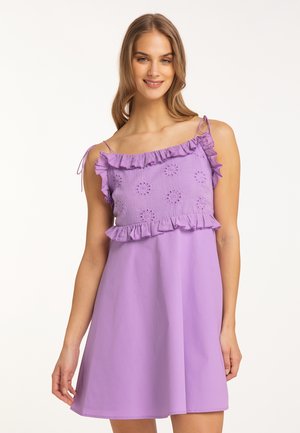 Junge Frau mit langen Haaren trägt ein ärmelloses violettes Kleid mit Rüschen und Lochmuster am Oberteil, lächelnd und vor hellem Hintergrund stehend.