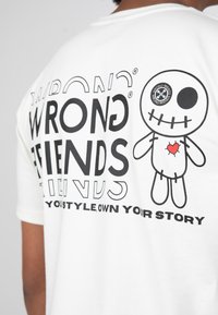 Bílý tričko s černým textem "WRONG FRIENDS" a karikaturním designem panenky se srdíčkem. Krátké rukávy, uvolněný střih.