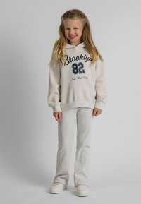 Lächelndes Mädchen mit langen blonden Haaren trägt einen beigefarbenen Hoodie mit dem Text Brooklyn 82, hellgraue Hose und weiße Sneaker und steht auf einem grauen Boden.