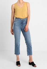 Donna che indossa un top senza maniche giallo senape a V, jeans chiari tagliati con orli sfilacciati e mule nere con tacco a blocco e punta aperta.