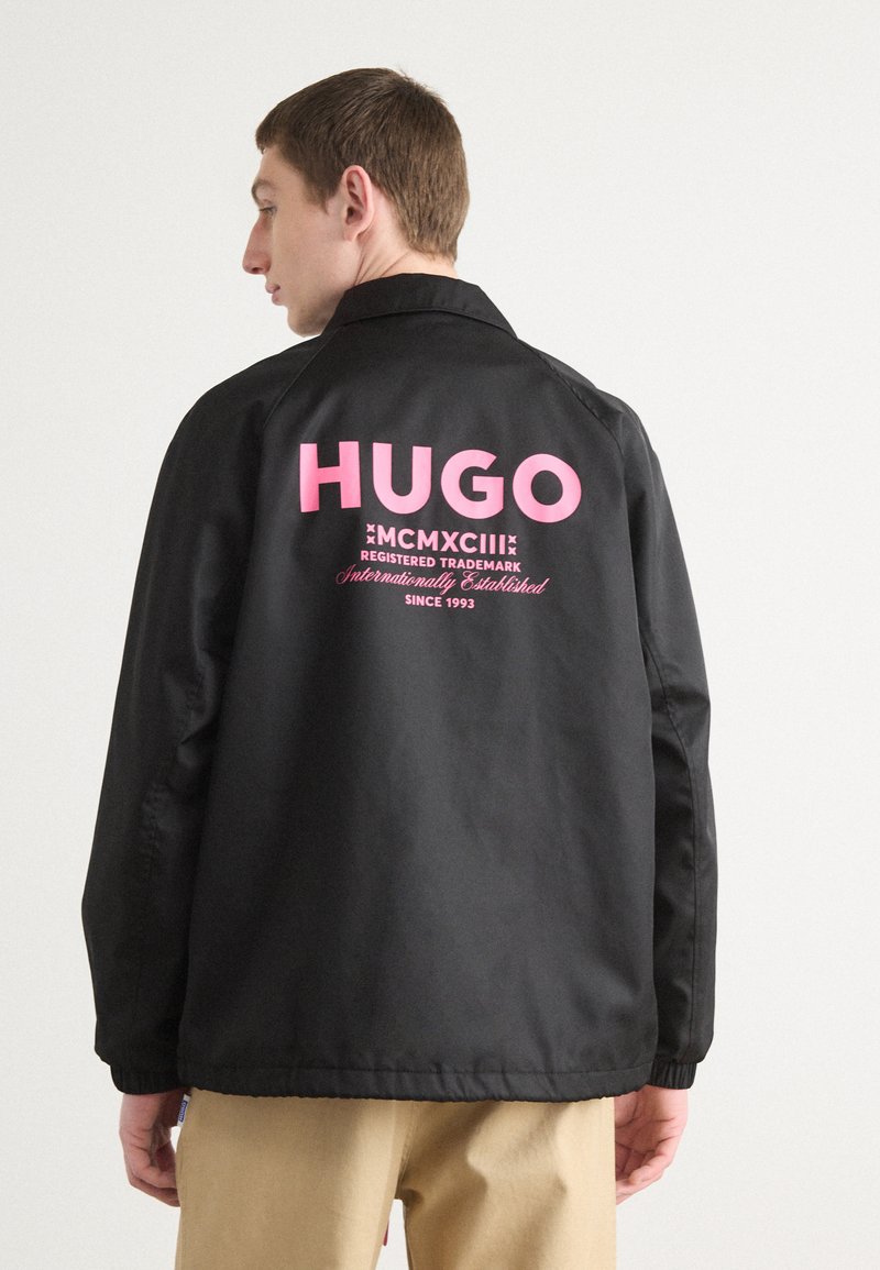 Črna bunda z rožnato "HUGO" logotipom na hrbtu, z gladko teksturo in standardnim ovratnikom. Očitne šive detajle ob šivih.