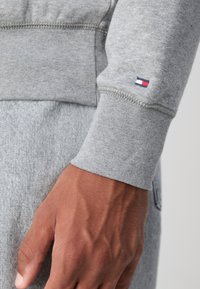Gros plan d'une main portant un sweat-shirt gris avec un petit logo Tommy Hilfiger rouge, blanc et bleu sur le poignet.