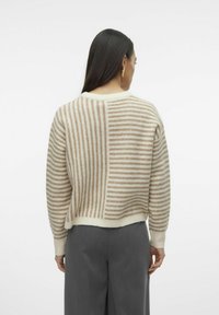 Creme- und beige gestreifter Strickpullover mit lockerer Passform, geripptem Halsausschnitt und Säumen, kombiniert mit grauen Hosen. Die Rückansicht zeigt ein asymmetrisches Muster.