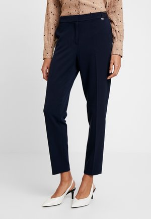 Kvinde iført marineblå skræddersyede ankelbukser, beige mønstret bluse og hvide slingback højhælede sko, stående mod en ensfarvet baggrund.