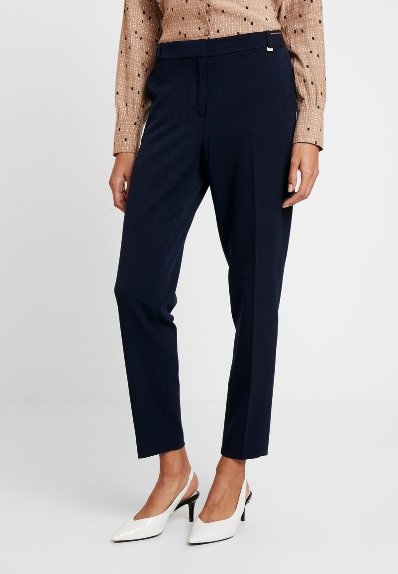 Femme portant un pantalon taille cheville bleu marine sur mesure, un chemisier beige à motifs et des escarpins blancs à bride arrière, debout devant un fond uni.