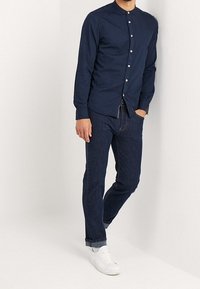 Mann in einem Hemd mit Marinebandkragen, dunklen blauen hochgekrempelten Jeans und weißen Sneakers, der mit einer Hand in der Tasche steht.