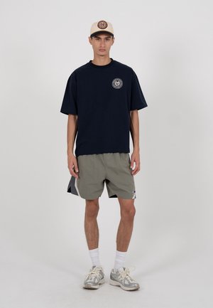 Mann mit beiger Kappe, schwarzem übergroßem T-Shirt mit Logo, grauer Sportshorts, weißen Socken und silbernen Turnschuhen vor einfachem Hintergrund stehend.