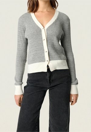 Femme portant un cardigan gris côtelé avec bordures et boutons blancs, associé à un jean noir taille haute à jambes larges, debout devant un fond uni.