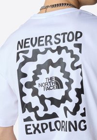 Biała koszulka z krótkim rękawem z czarnym abstrakcyjnym wzorem graficznym i napisem "NIGDY NIE PRZESTAWAJ BADAĆ" z logo "THE NORTH FACE".