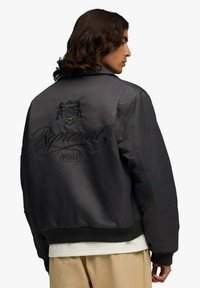 Giacca bomber nera in materiale liscio, con logo e testo in rilievo sulla schiena. Polsini e orlo a coste aggiungono texture e aderenza.