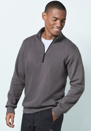 Gray zip-up sweatshirt with a high collar, ribbed cuffs, and a logo patch on the hem. Worn with dark pants, featuring a smooth texture.