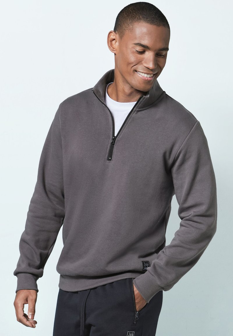 Gray zip-up sweatshirt with a high collar, ribbed cuffs, and a logo patch on the hem. Worn with dark pants, featuring a smooth texture.