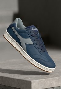 Navy blue sneakers made of suede and synthetic materials, featuring a gray logo, textured upper, and white rubber sole with a brown accent.