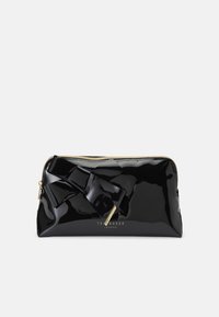 Bolso de charol negro con un acabado brillante, con un destacado detalle de lazo y hardware en tono dorado. Cierre de cremallera en la parte superior.