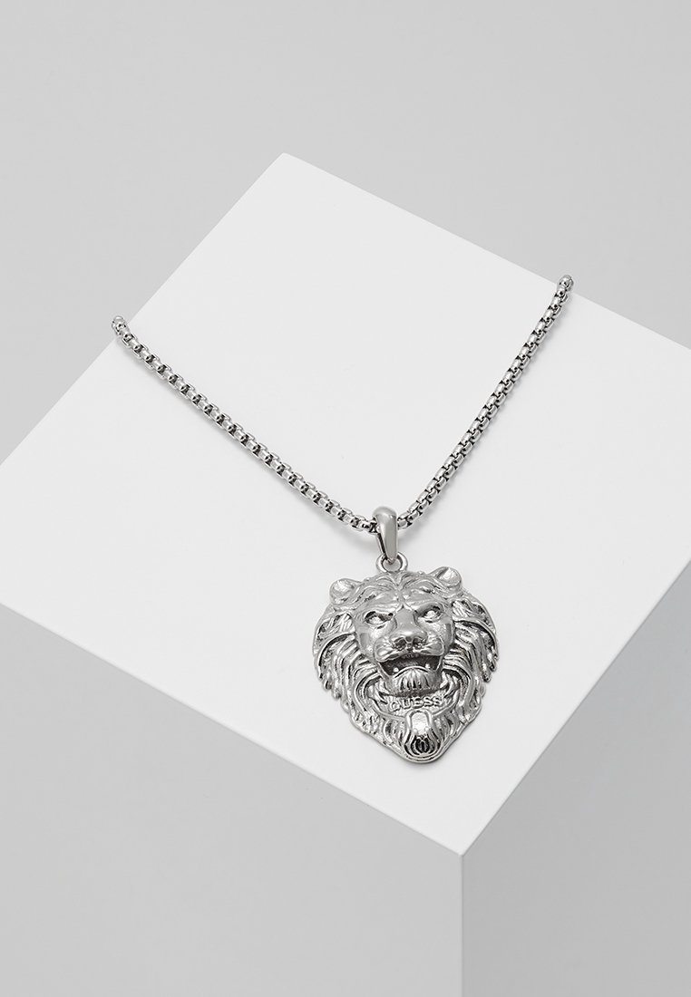 Collier en chaîne argentée avec un pendentif tête de lion détaillé gravé du mot "Guess" sur une surface blanche inclinée.