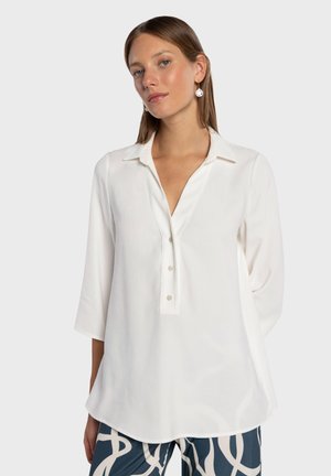 Blouse blanche à manches longues avec col, encolure en V et fermeture à boutons sur le devant. Texture lisse, coupe légèrement ample et ourlet arrondi.