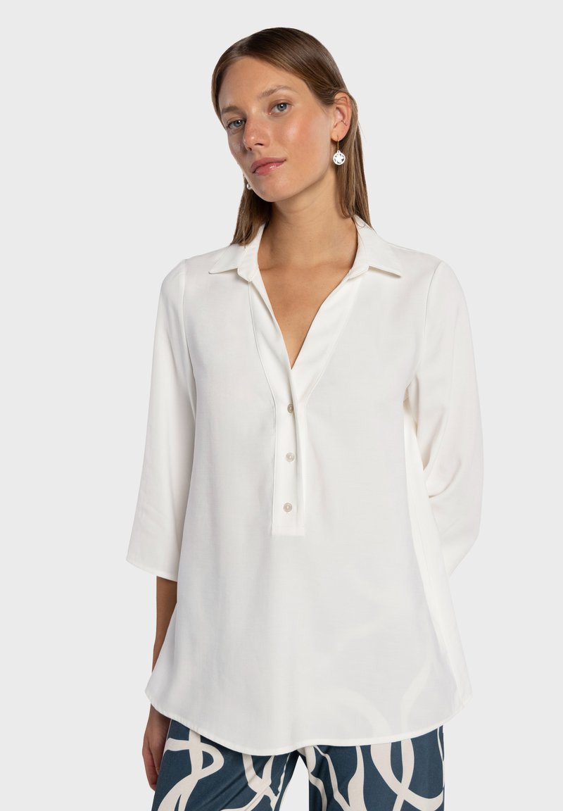 Blouse blanche à manches longues avec col, encolure en V et fermeture à boutons sur le devant. Texture lisse, coupe légèrement ample et ourlet arrondi.