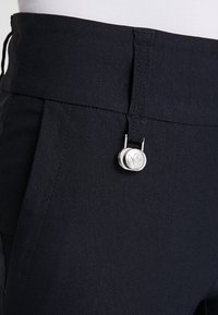 Un pantalon noir en tissu léger avec une fermeture à bouton dotée d'un détail circulaire en argent et une taille lisse et texturée.