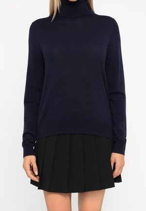 Femme portant un pull à col roulé bleu marine foncé et une jupe plissée noire, vue des épaules jusqu’au milieu des cuisses, sur fond blanc.