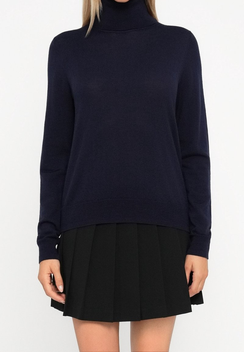 Femme portant un pull à col roulé bleu marine foncé et une jupe plissée noire, vue des épaules jusqu’au milieu des cuisses, sur fond blanc.