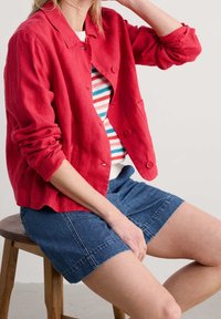 Red linen button-up jacket with rolled sleeves, paired with a striped sweater and denim shorts. Wood stool in a neutral setting.