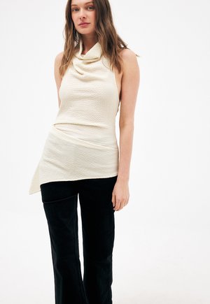 Femme portant un haut crème sans manches à texture avec un col drapé et un ourlet asymétrique, associée à un pantalon noir, debout devant un fond blanc.