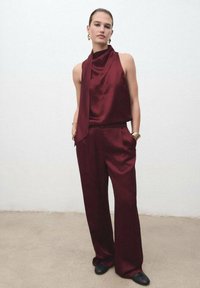 Top de satén burdeos sin mangas con un detalle de bufanda drapeada, combinado con pantalones holgados; textura suave, diseño minimalista y bolsillos laterales.