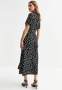 Vestido midi negro con lunares, mangas cortas, una cintura ajustada y una falda fluida. El material parece suave y ligero. Sandalias negras con tiras.