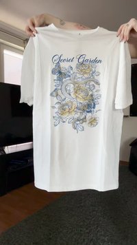 Hvid T-shirt holdt op, der viser et "Secret Garden" design med gule roser, en slange og blå sommerfugle omgivet af blade.